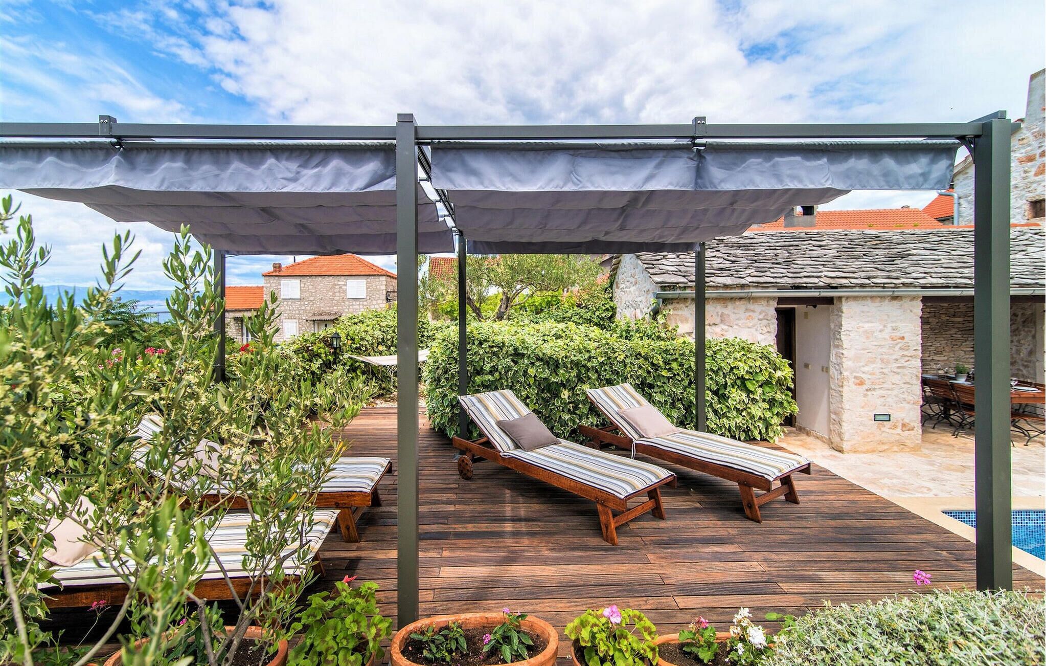 Een houten terras met twee lounge fauteuils onder een pergola, omringd door potplanten en een stenen gebouw op de achtergrond.
