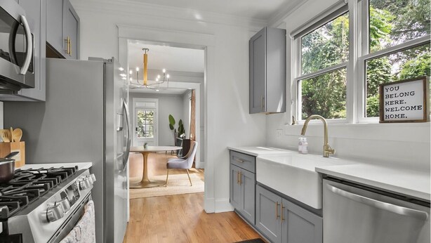 A modern kitchen with grey cabinets, a stainless steel refrigerator, and a stove. There is a window with a view of trees and a sign that says "YOU BELONG HERE. WELCOME HOME." - Airport Cottage  (East Point)