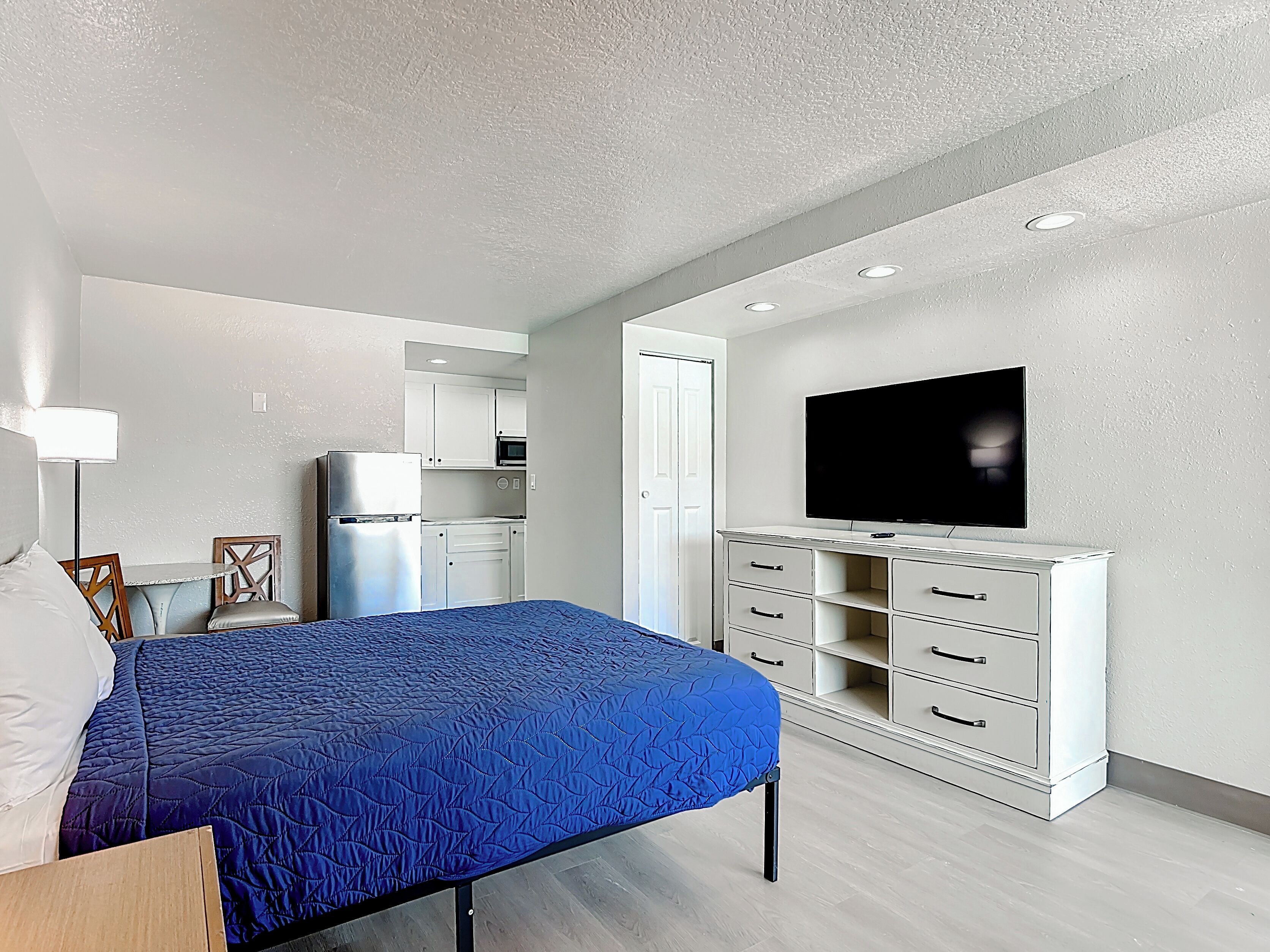 A bedroom with a bed, a TV, a kitchenette, and a door leading to another room.