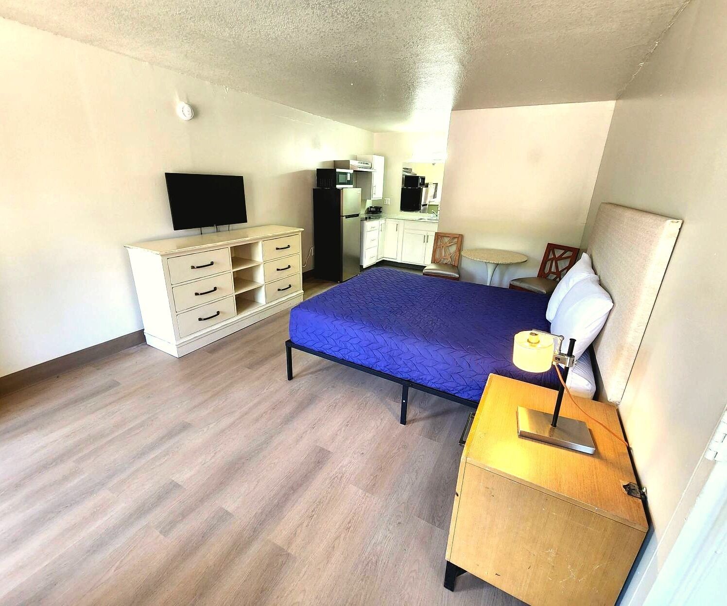 Un dormitorio con cama, cómoda, escritorio con lámpara, televisor, microondas y una mesita.