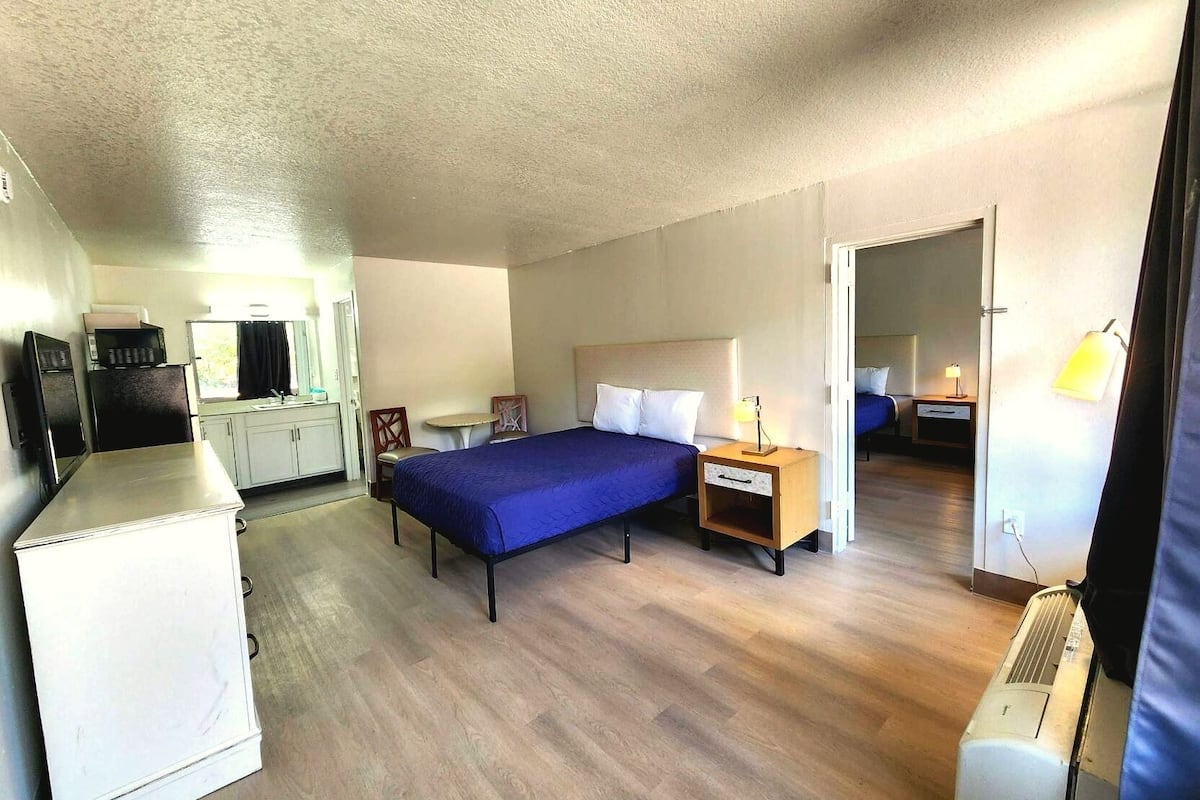 A compact hotel room with a bed, bedside table, lamp, and kitchenette.