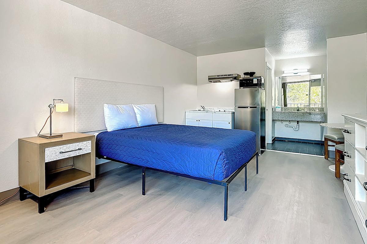 A bedroom with a bed, bedside table, lamp, and a kitchenette area with a refrigerator and microwave.