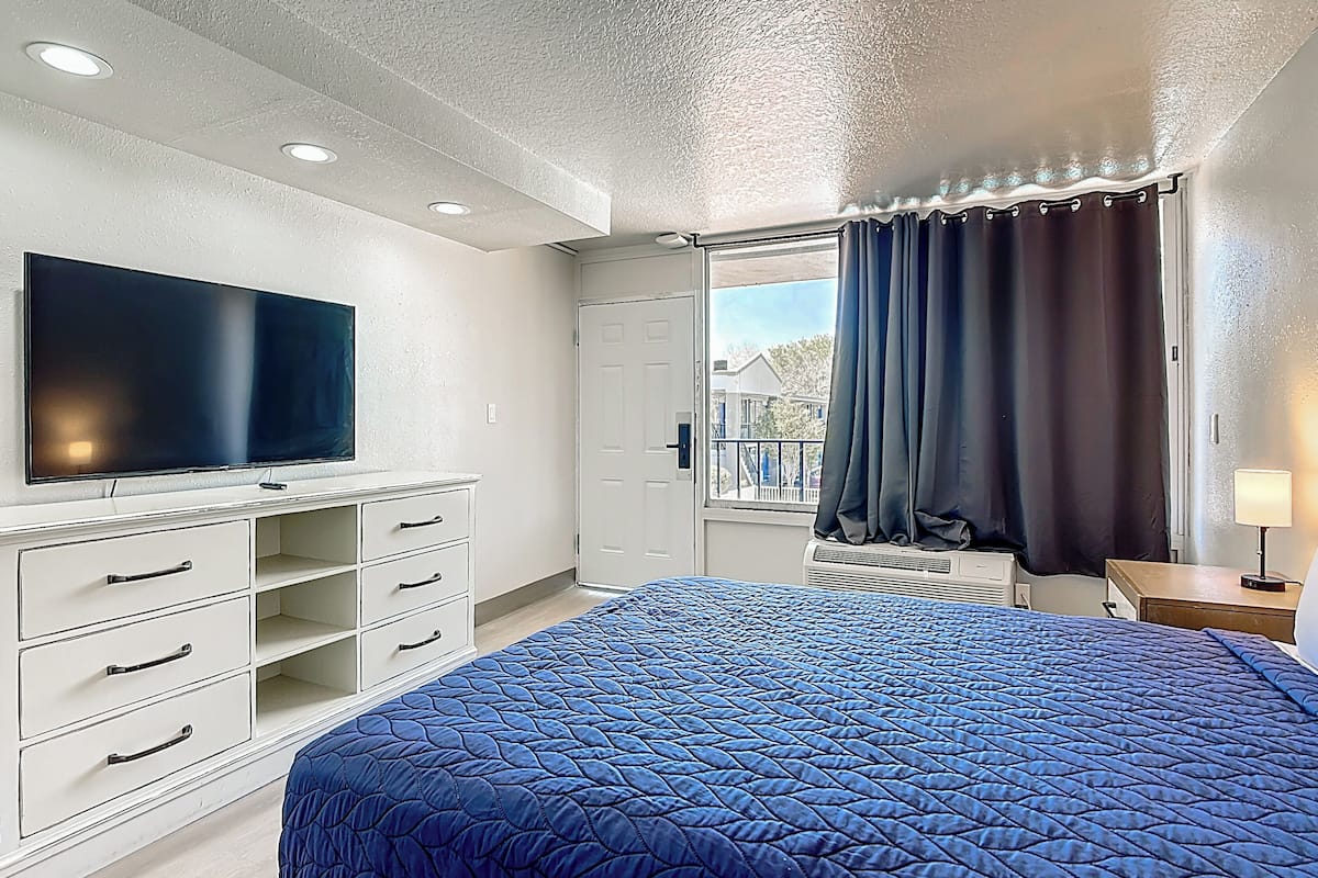 A bedroom with a bed, a TV, a dresser, a window with curtains, and a view of buildings outside.