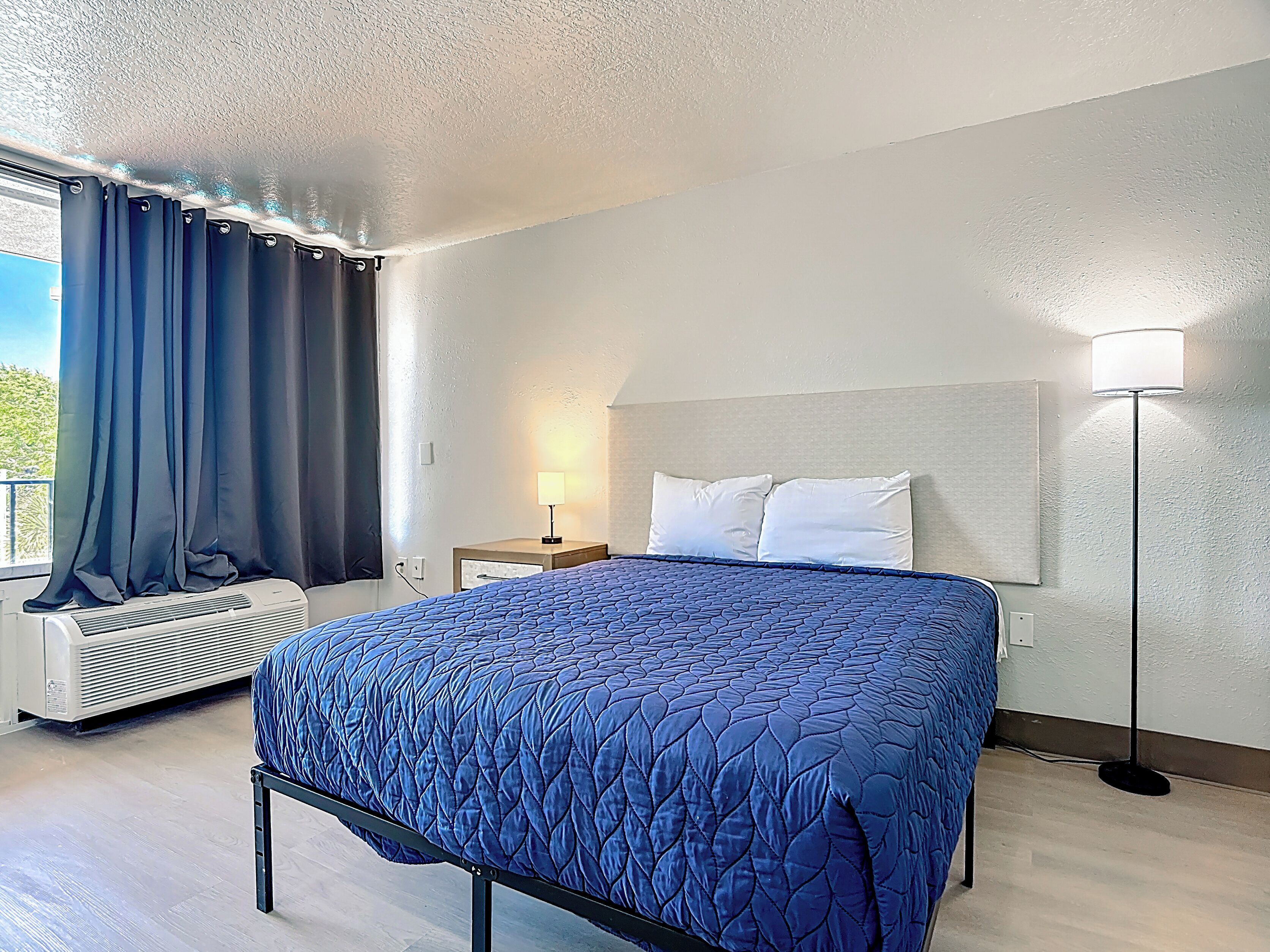 A hotel room with a bed, a nightstand, a lamp, an air conditioning unit, and a window with curtains.