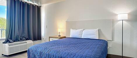 A hotel room with a bed, a nightstand, a lamp, an air conditioning unit, and a window with curtains.