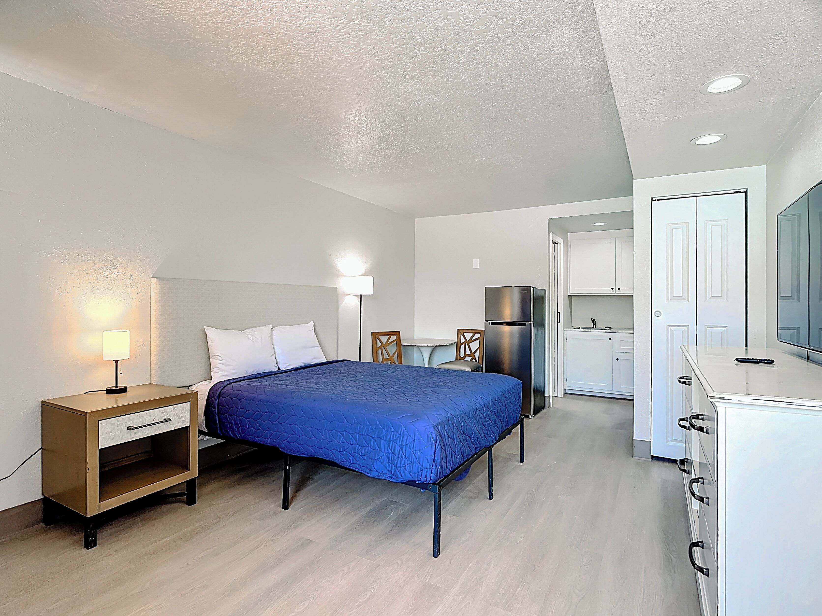 A bedroom with a bed, bedside table, lamp, and a kitchenette area with a refrigerator and cabinets.