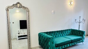 A room with a teal velvet sofa, a large ornate mirror, a white wall, a small white bench, and a wall-mounted light fixture. - JABEEN'S HOUSE (Bezons)