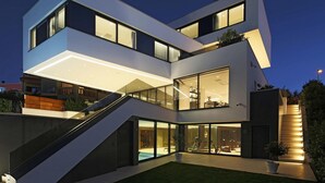 Una moderna casa a più piani con facciata in vetro, area salotto esterna e una scalinata che conduce ai piani superiori.