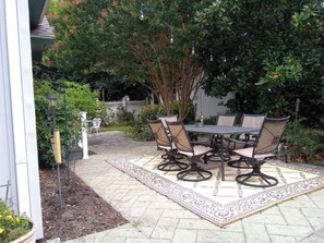 An outdoor patio with a round table and chairs, surrounded by a patterned rug, under a tree.