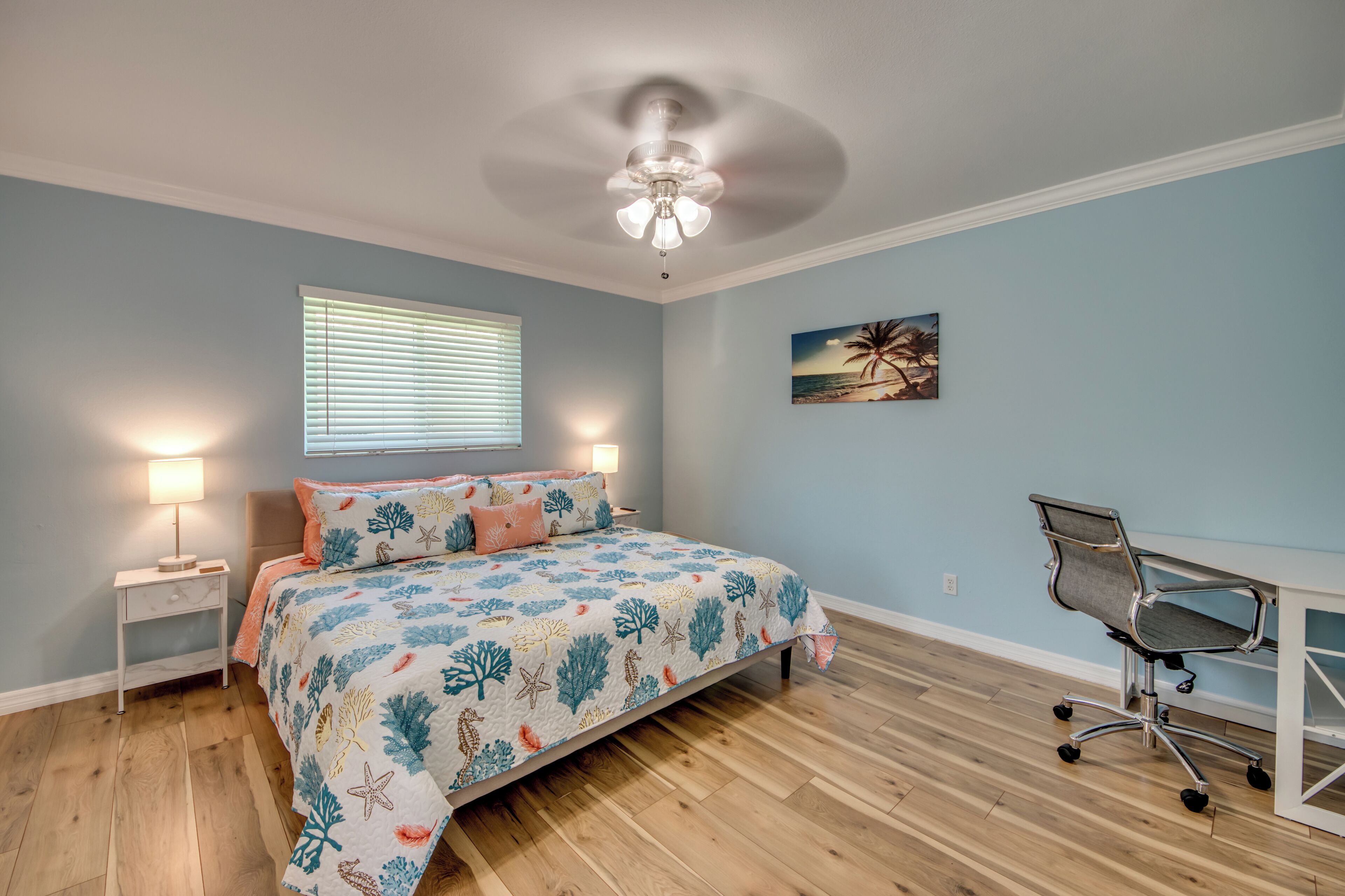 A bedroom with a bed, a desk, a chair, a ceiling fan, a nightstand, and a window with blinds.
