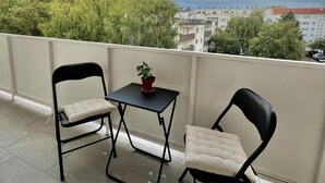 A balcony with two black chairs and a small table, a potted plant, and a view of buildings and trees under a partly cloudy sky.