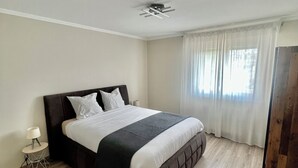 A modern bedroom with a large bed, a bedside table with a lamp, and a window with sheer curtains.