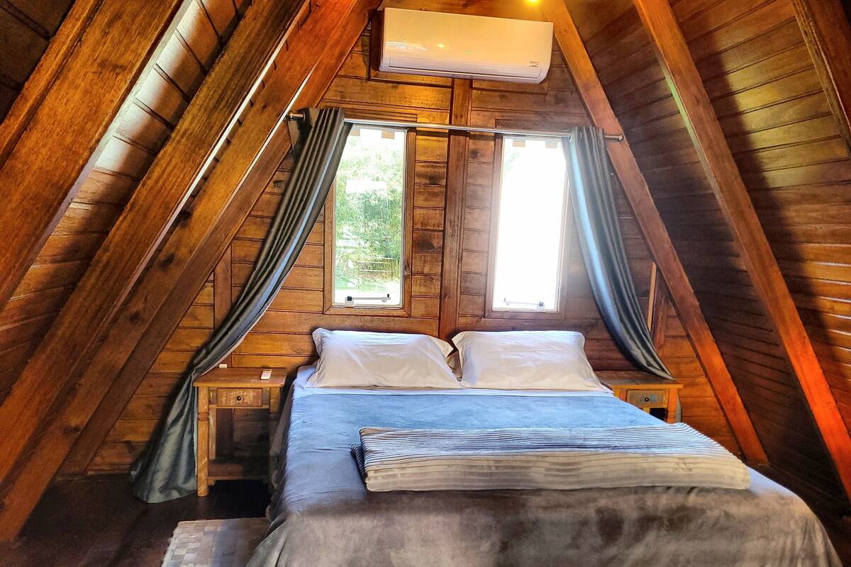 Un dormitorio acogedor en el ático con techo inclinado de madera, una cama con ropa blanca y una ventana con cortinas.