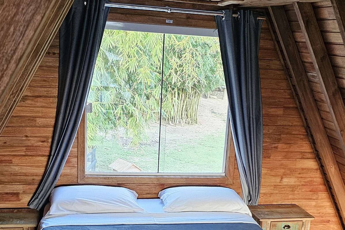Un dormitorio de cabaña de madera con cama, cortinas y ventana con vistas a follaje.
