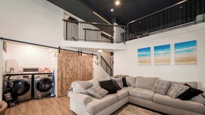 A modern living room with a grey sectional sofa, a washing machine, and a staircase leading to an upper level.