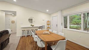 Un espace repas avec une table en bois et des chaises blanches, un comptoir blanc avec des tabourets, et une vue sur la verdure par une fenêtre.