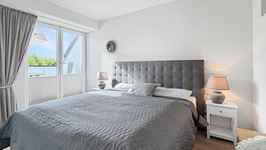 A bedroom with a large bed, a nightstand, a lamp, a window with curtains, and a wall decoration. - 3002 - Grömitz - Wohnung 3002 am Schoor in Grömitz (Grömitz)