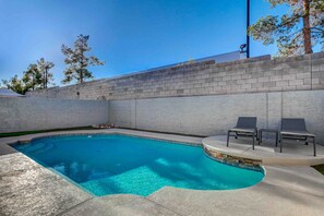 A swimming pool with a curved edge, surrounded by a brick wall, and two lounge chairs on a raised platform.