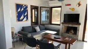 Un salon moderne avec un canapé gris, une table à manger en bois accompagnée de deux chaises, un téléviseur à écran plat fixé au-dessus d’une cheminée carrelée, et une grande fenêtre donnant sur l’extérieur.