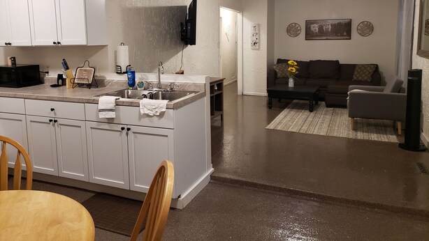 Eine moderne Küche mit weißen Schränken, einer eingebauten Mikrowelle und einem Spülbecken. Ein Essbereich mit einem Holztisch und Stühlen lädt zum Verweilen ein. Das Wohnzimmer bietet ein dunkles Sofa, einen Couchtisch und einen an der Wand befestigten Fernseher.