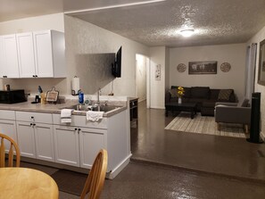Eine moderne Küche mit weißen Schränken, einer eingebauten Mikrowelle und einem Spülbecken. Ein Essbereich mit einem Holztisch und Stühlen lädt zum Verweilen ein. Das Wohnzimmer bietet ein dunkles Sofa, einen Couchtisch und einen an der Wand befestigten Fernseher.