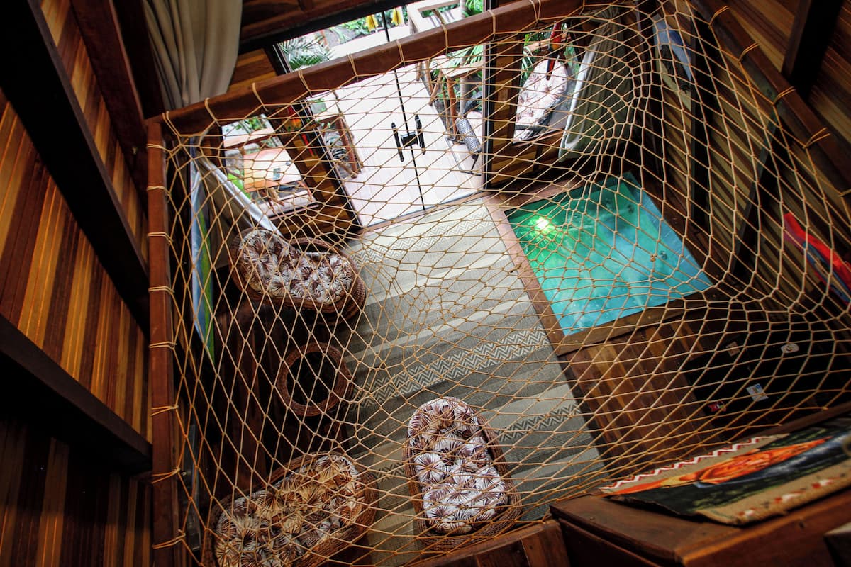 A room with a wooden floor, a bed with a patterned bedspread, a small table with a book, and a window with curtains.