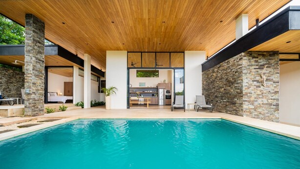 Un área de piscina moderna con techo de madera, pilares de piedra y una cocina al fondo.