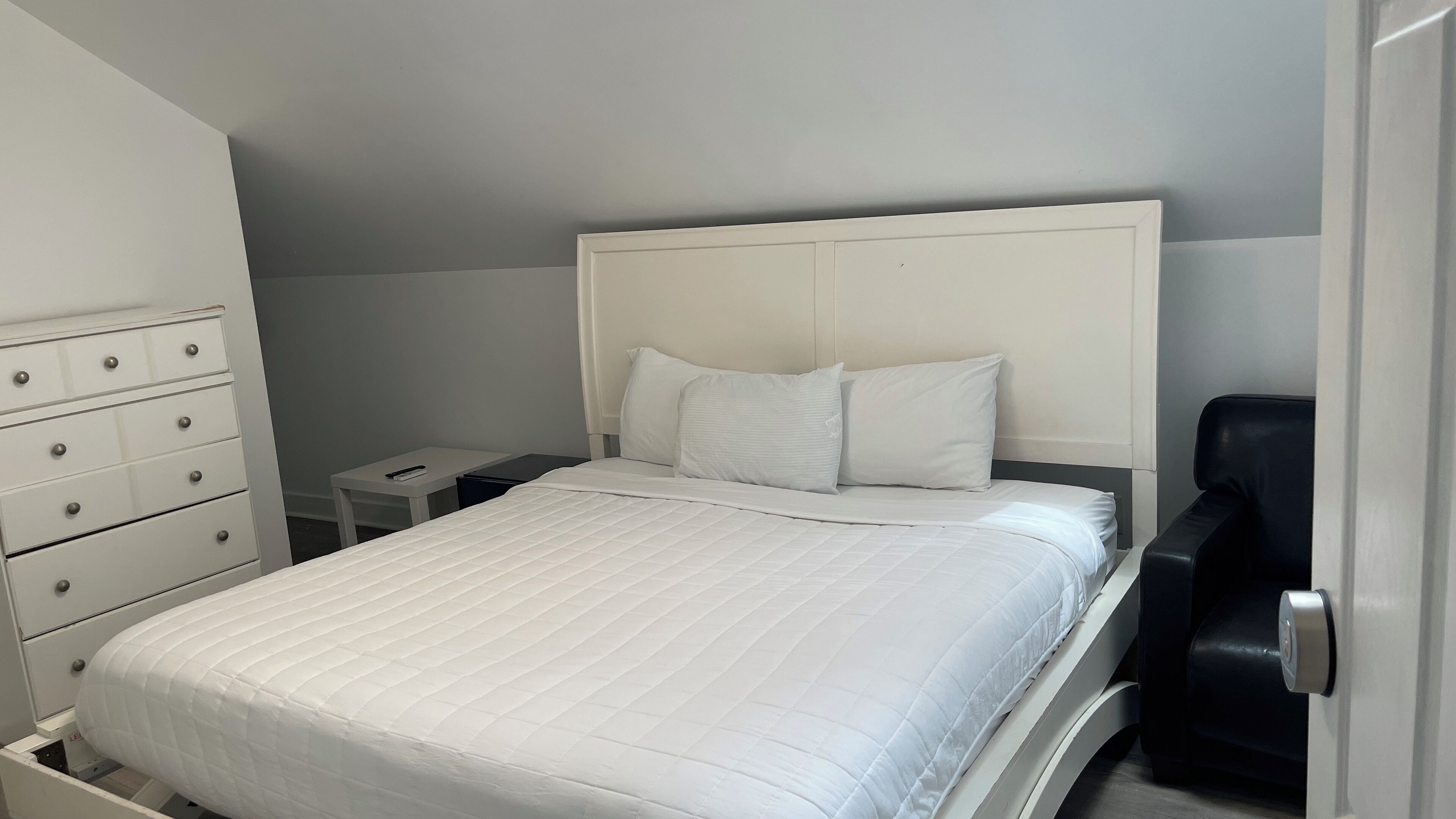A neatly made bed with a white headboard and two pillows, a white dresser with drawers, a bedside table with a lamp, and a black chair.