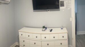 Une pièce avec une commode blanche, un téléviseur à écran plat fixé au mur, une petite plante en pot et une bouteille d’eau posée sur la commode.