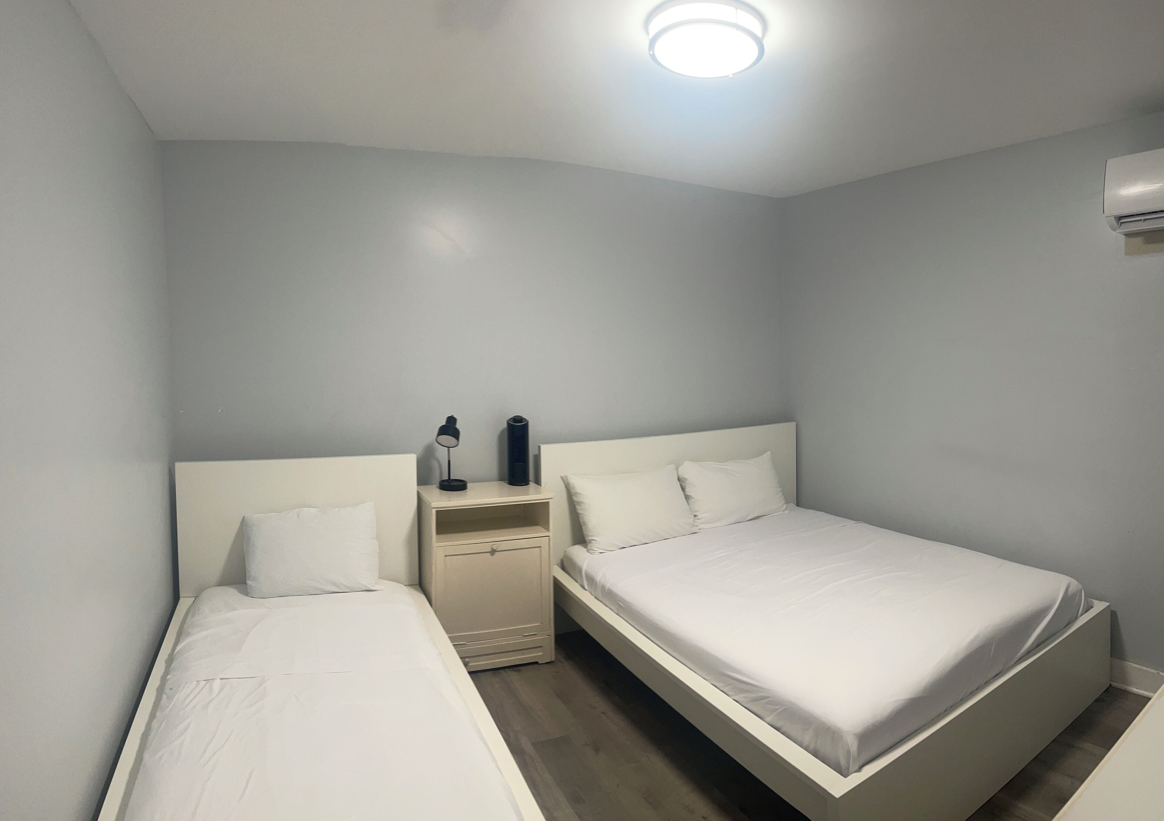 Ein kleines, sauberes Zimmer mit zwei Einzelbetten, einem Nachttisch mit Lampe und Deckenleuchte.