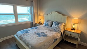 A bedroom with a bed, two bedside tables with lamps, a ceiling fan, and a view of the ocean through a window. - 1/1 Ocean View Condo at Sandpiper. Watch rocket launches from the balcony! (Satellite Beach)
