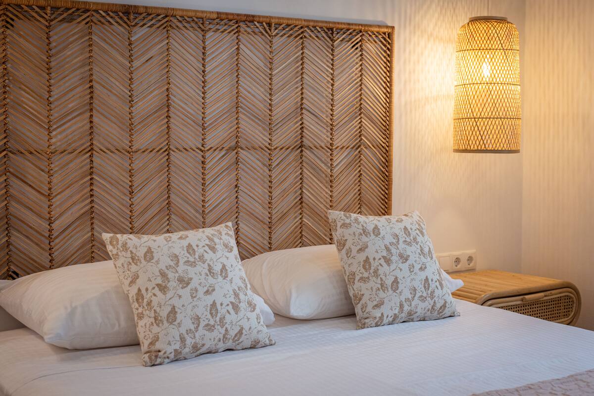 A bed with white linens and two patterned pillows, a woven headboard, and a wall-mounted light fixture.