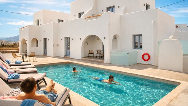 Un espace aménagé au bord de la piscine, avec des chaises longues, une piscine et un bâtiment aux murs blancs et aux arcades.