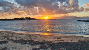 Une plage avec un coucher de soleil sur l’eau, une côte lointaine parsemée de bâtiments et un ciel nuageux.