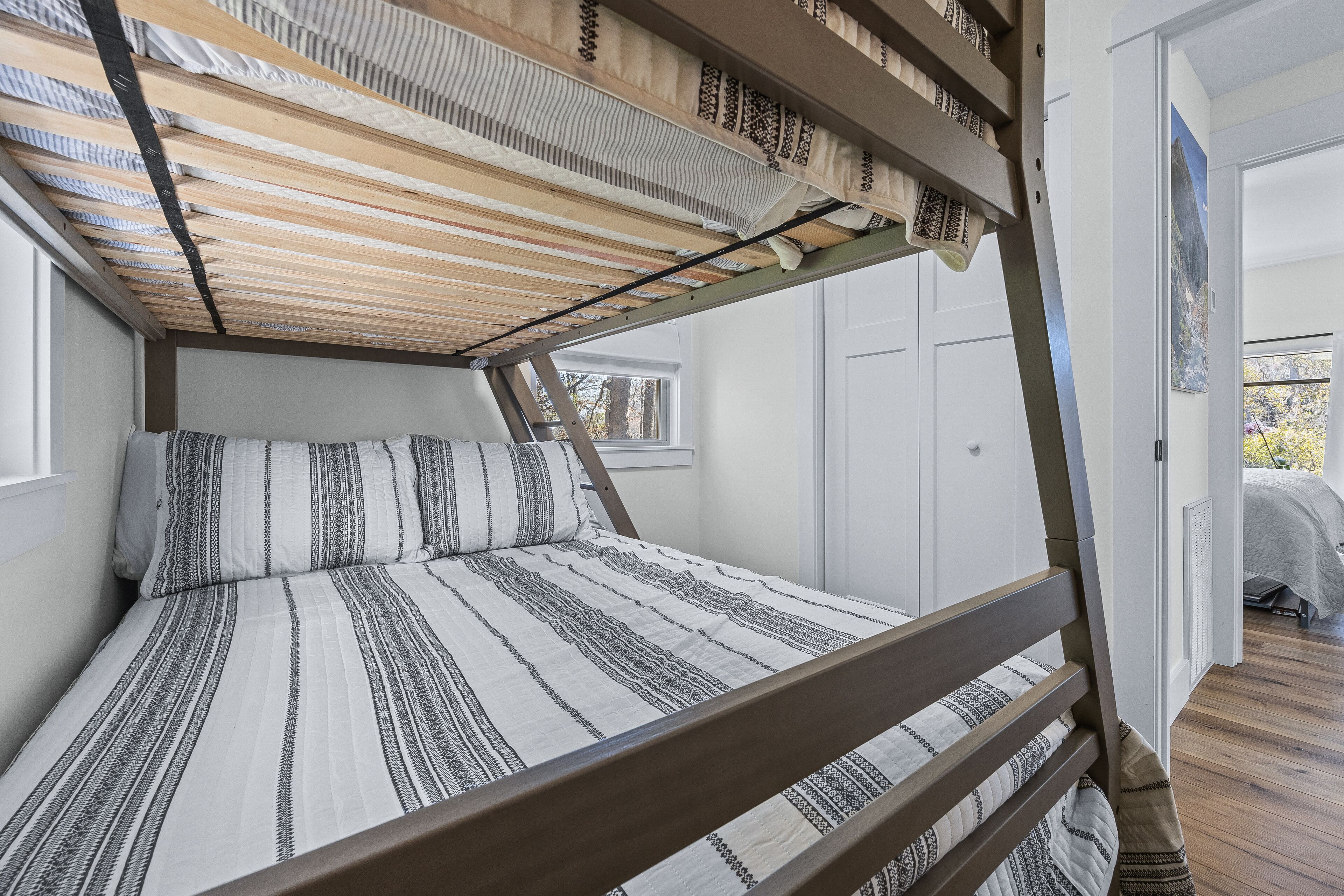 A bunk bed with a striped bedspread, a wooden headboard, and a window with a view of trees.
