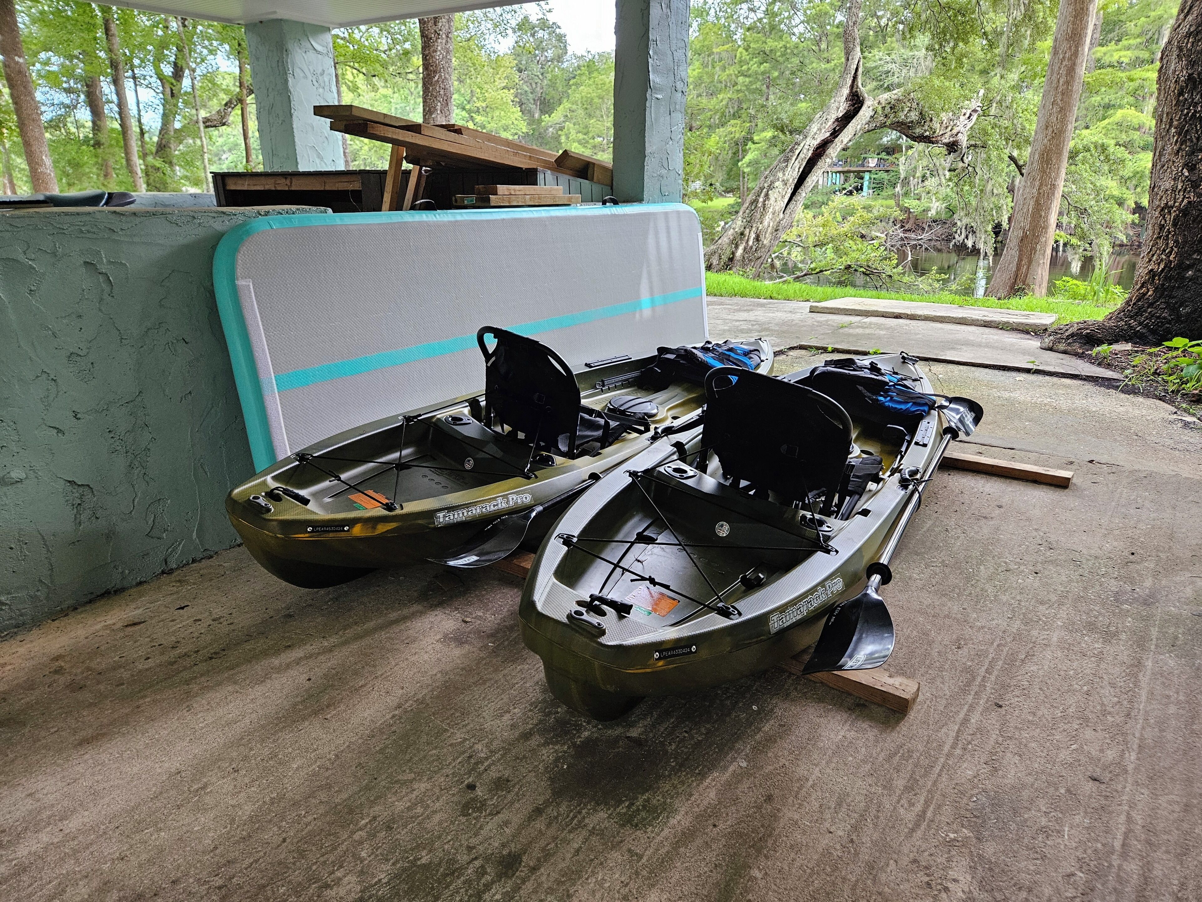 Two boats, a kayak, and a canoe, docked at a covered area with a concrete floor and a view of a forested area.