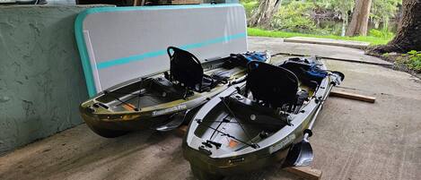 Two boats, a kayak, and a canoe, docked at a covered area with a concrete floor and a view of a forested area.