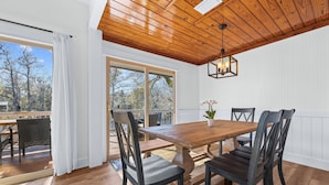 A dining area with a wooden table and chairs, large windows, and a view of trees.