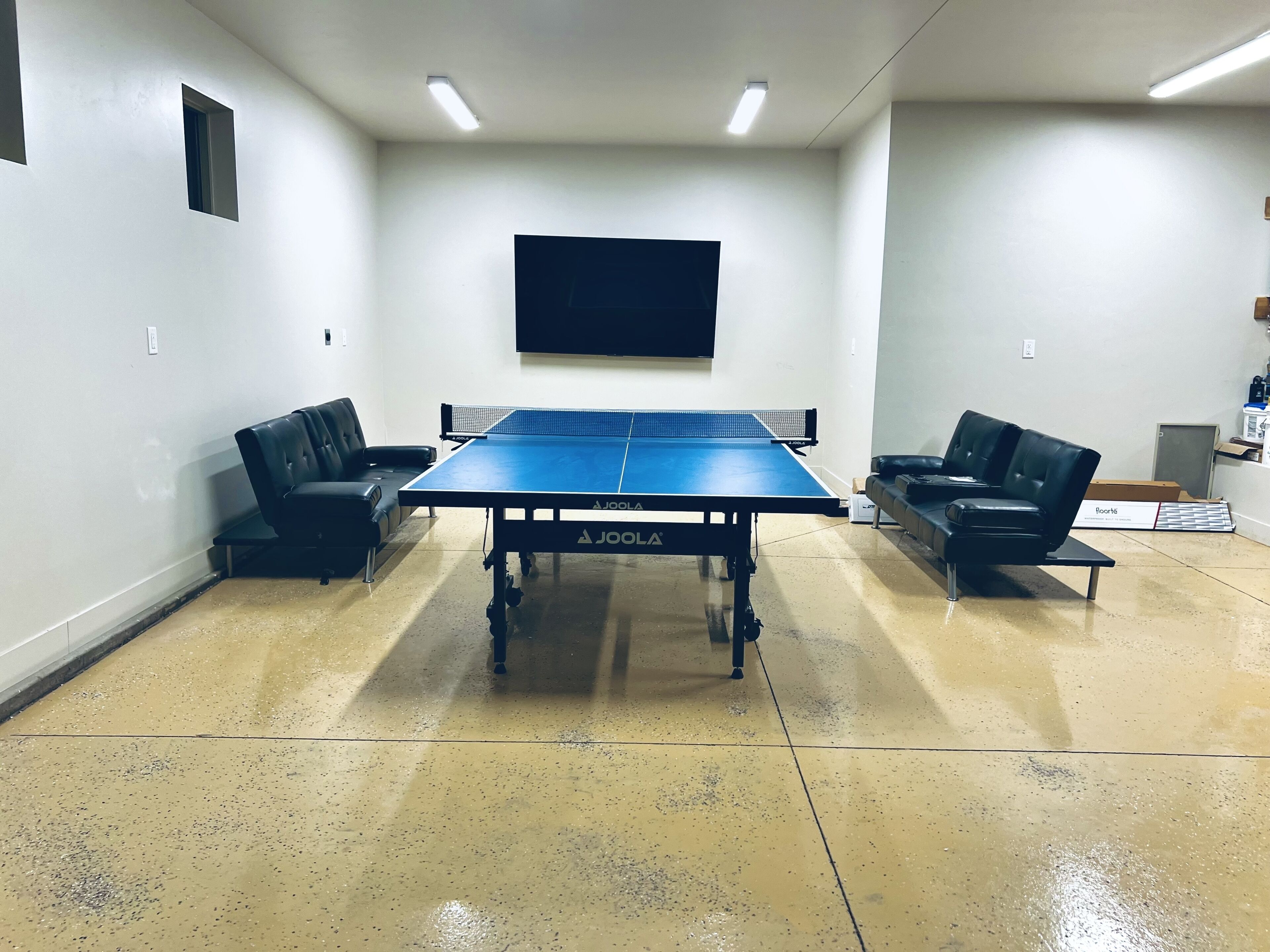 A ping pong table in a room with white walls, two black leather chairs, and a mounted television.