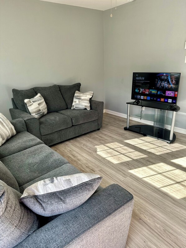 A modern living room with a grey sofa, a glass coffee table, and a flat-screen TV displaying a streaming service interface.