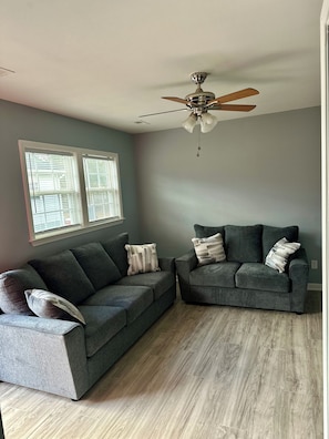 A living room with two grey sofas, a ceiling fan, and a window with blinds.