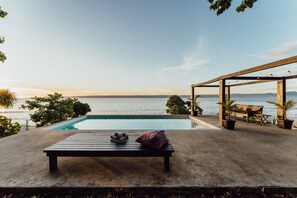 Un espace aménagé au bord de la piscine, comprenant un banc en bois, une petite table avec une serviette et un coussin, ainsi qu’une pergola en bois avec un banc et des plantes en pot.