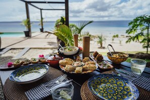 Une table à manger dressée avec différents plats, donnant sur la plage.
