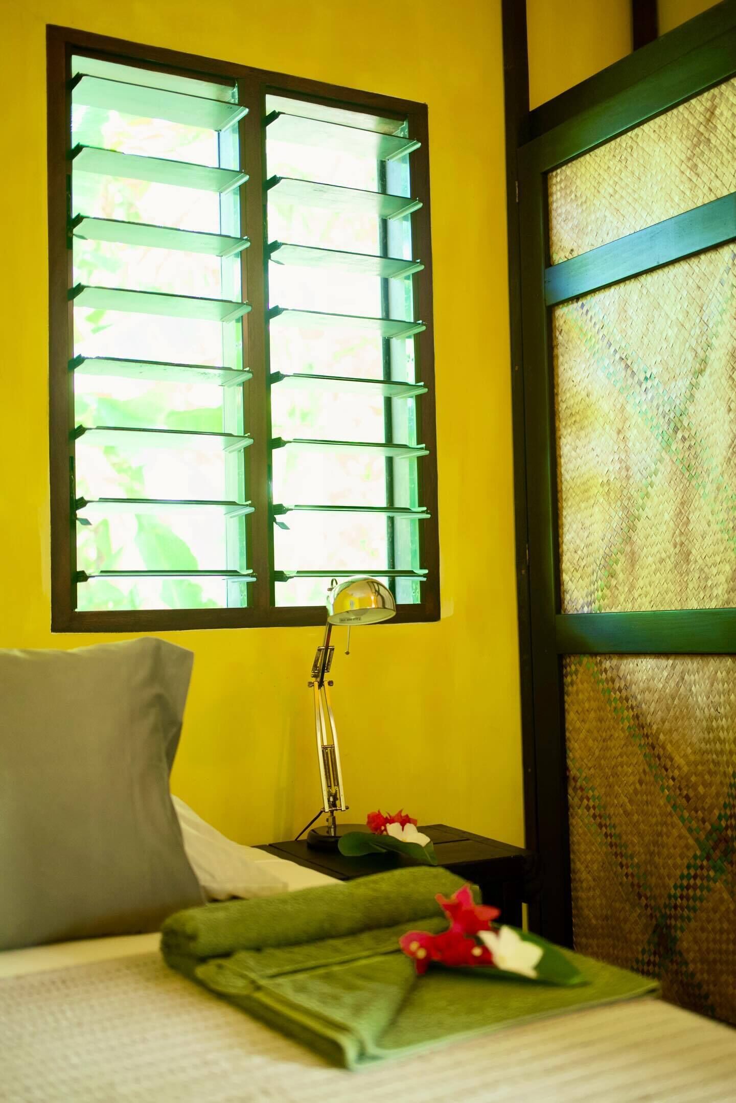 A bed with a green blanket, a pillow, a bedside lamp, and a window with blinds.
