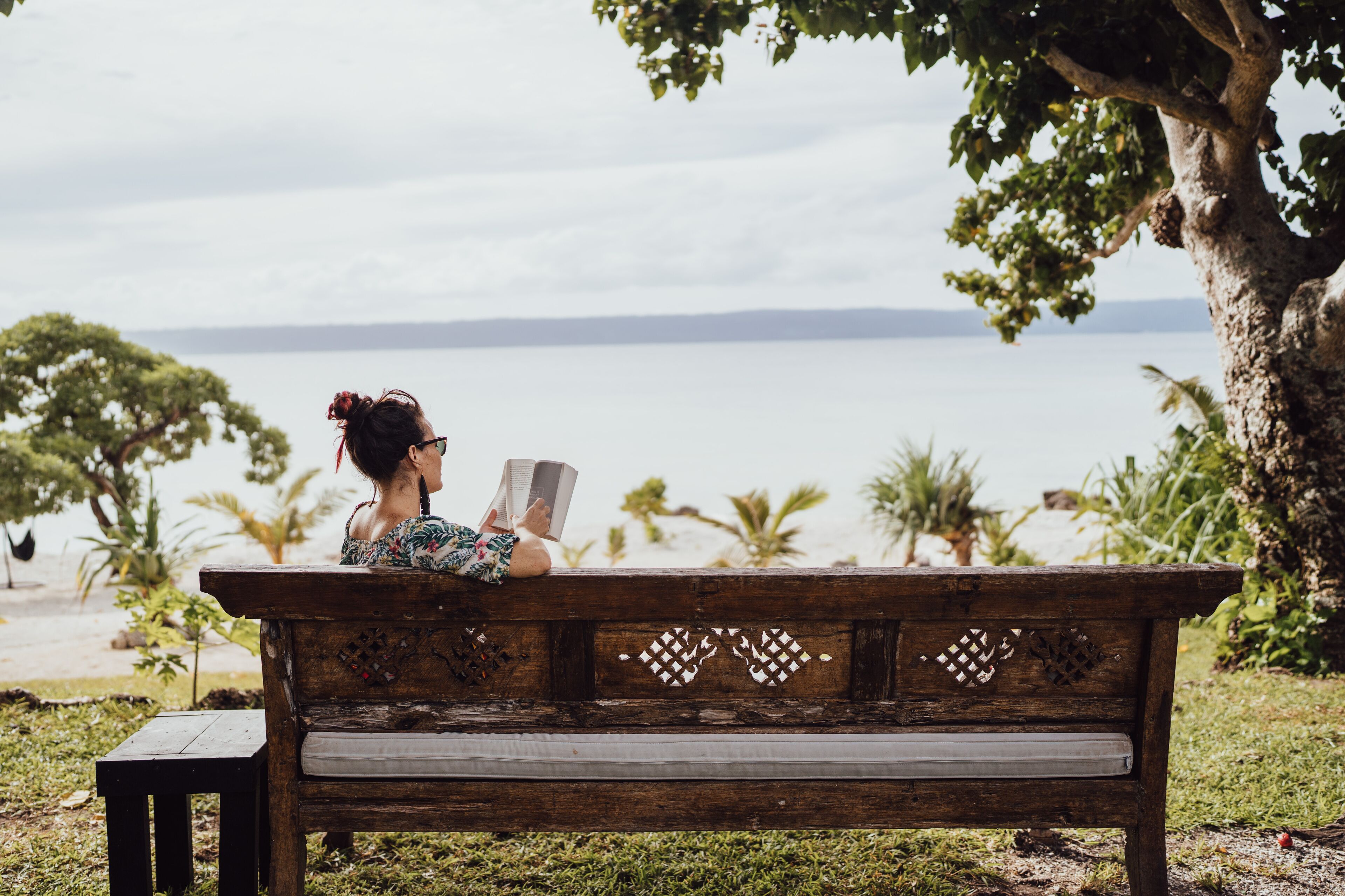 A wooden bench with intricate carvings, situated outdoors with a view of the sea and palm trees in the background.