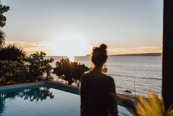 Une personne se tient près d’une piscine, avec une vue sur le coucher de soleil qui se reflète sur l’eau.