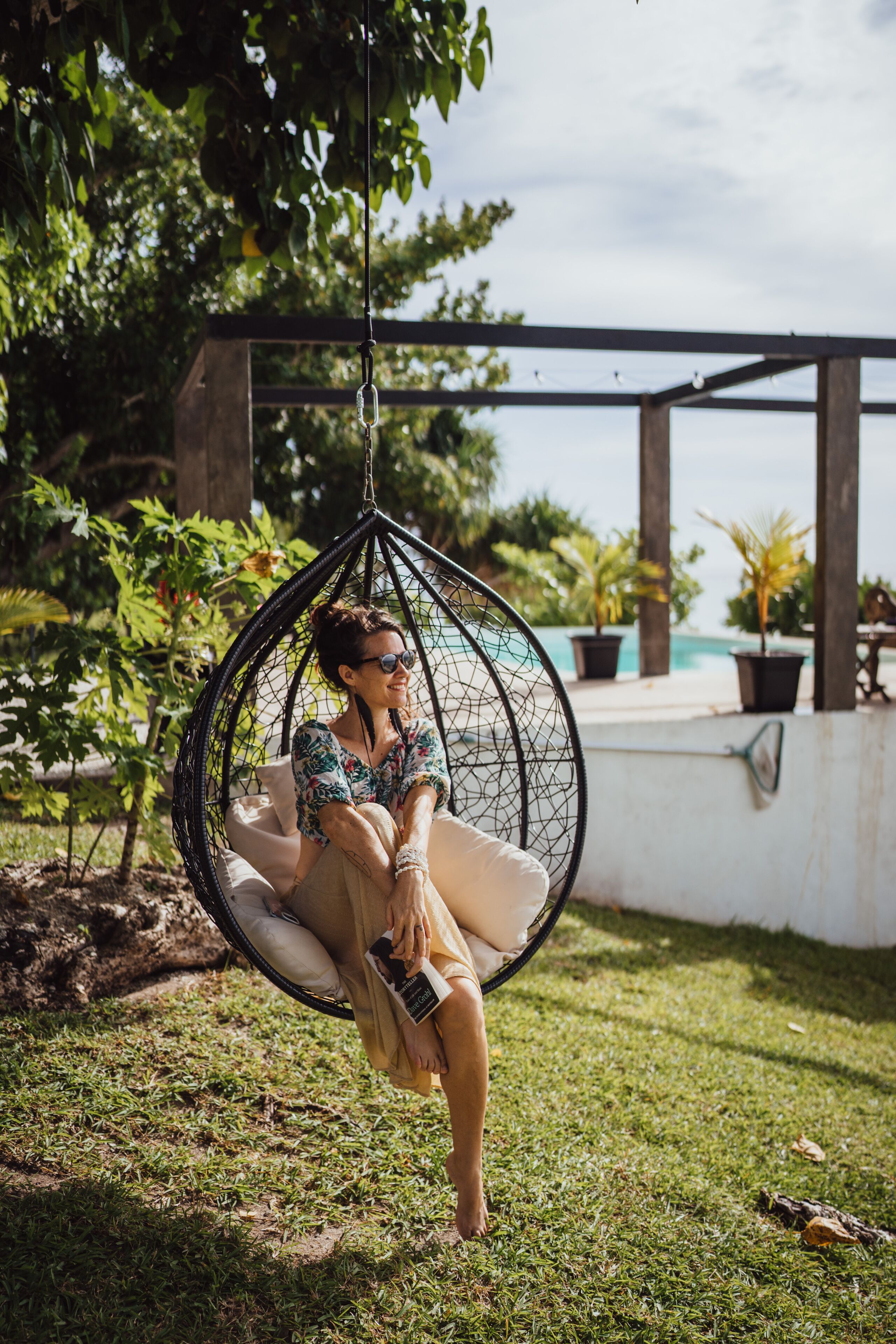 A person sitting in a hanging chair in a garden area with a pool in the background.
