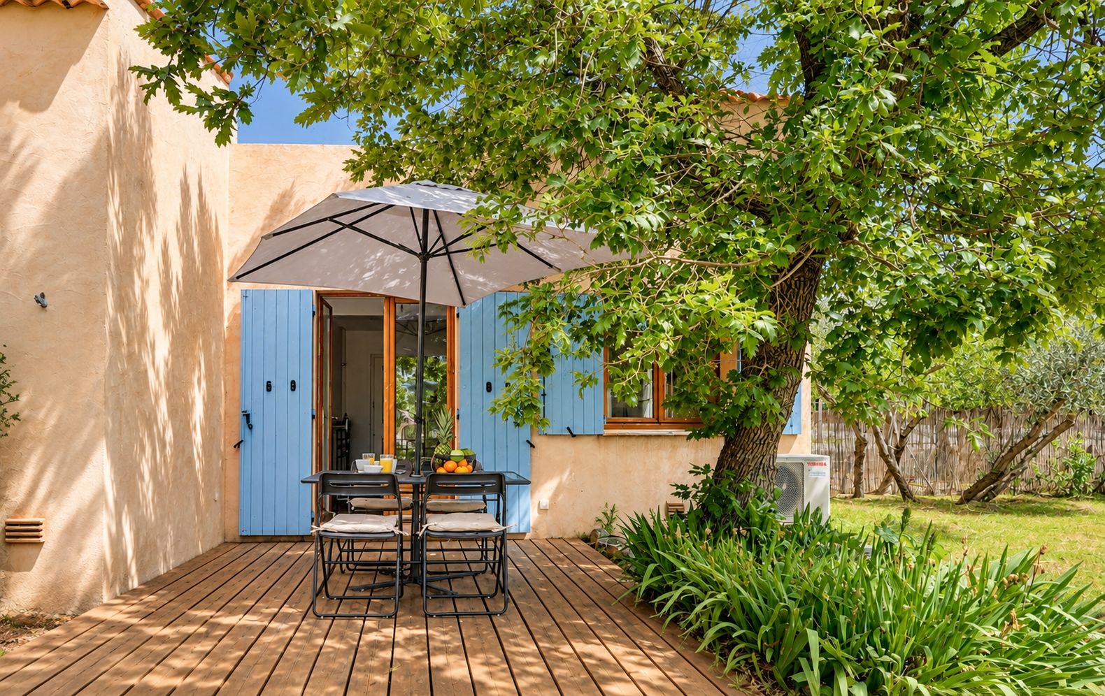 A patio with a table and chairs, an umbrella, and a tree providing shade.