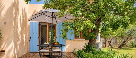 A patio with a table and chairs, an umbrella, and a tree providing shade.
