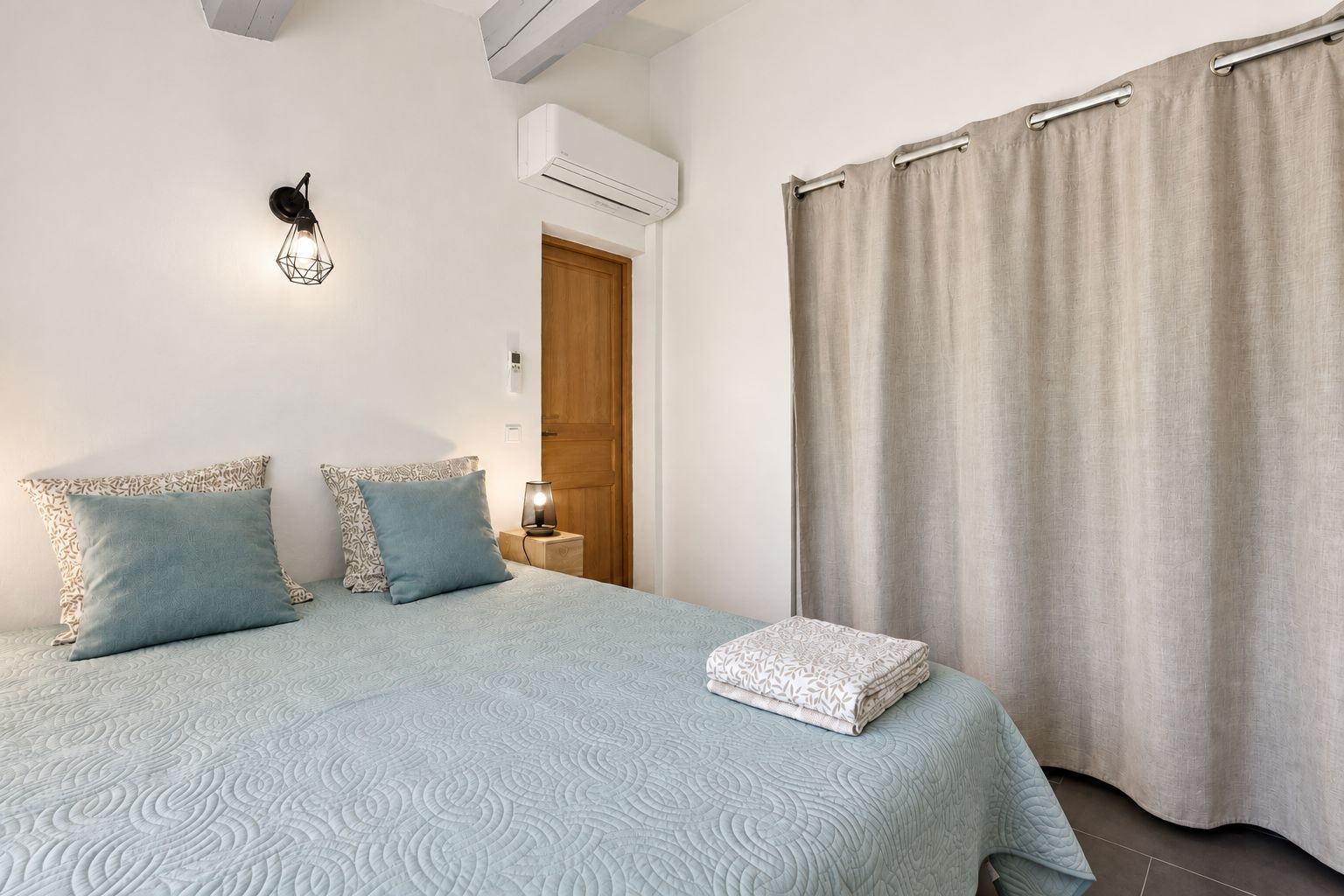 Een slaapkamer met een bed, kussens, een bedlampje, een airconditioningapparaat, een deur en een gordijn.